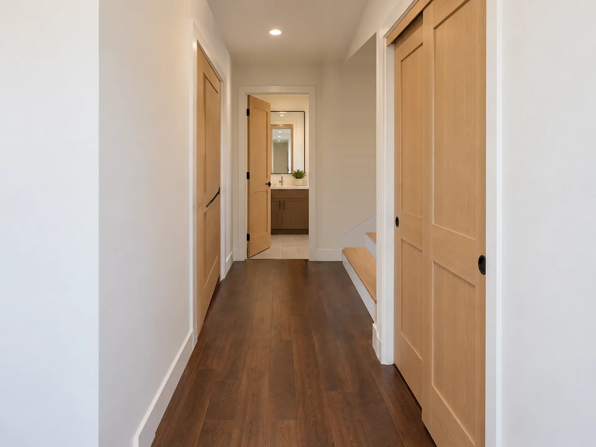 Rendering of the ADU 1200-2 hall: a dark plank floor between white walls, the closed light-oak door to bedroom 3 near the end on the left, the bath door open at the end with a walnut vanity and mirror beyond, a coat closet’s light-oak sliding doors on the right, and past them the first oak steps of the stair, climbing to the right behind the closet.