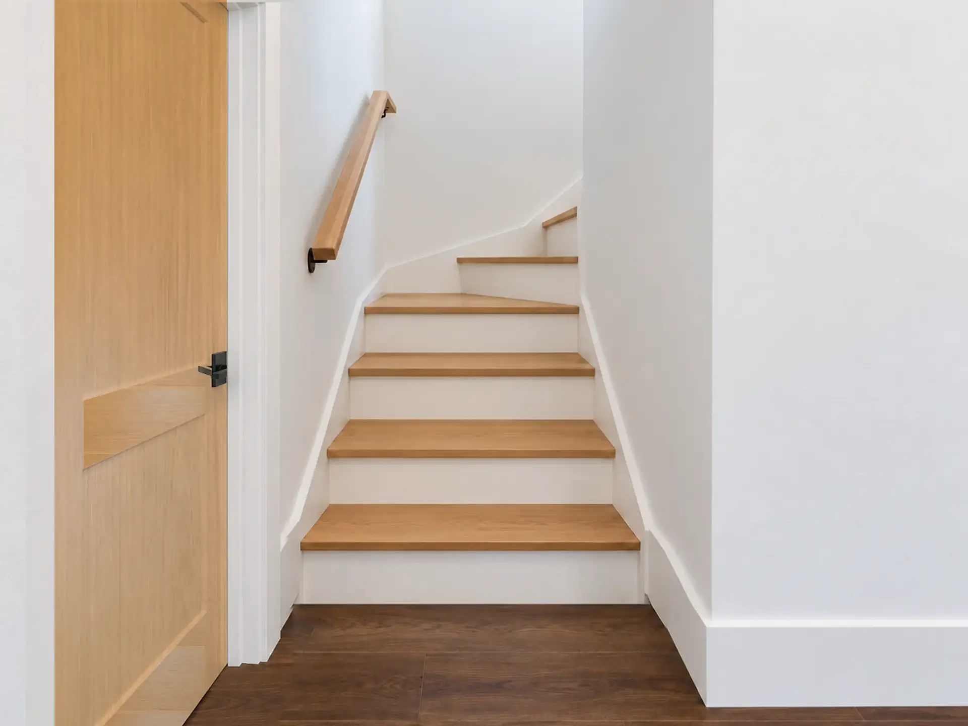 Rendering of the ADU 1200-2 stair from the hall: oak treads on white risers climbing between white walls and winding to the right at the top, a light-oak handrail on the left-hand wall, the closed light-oak door of the hall bath at the left edge, and the hall’s dark plank floor in front.