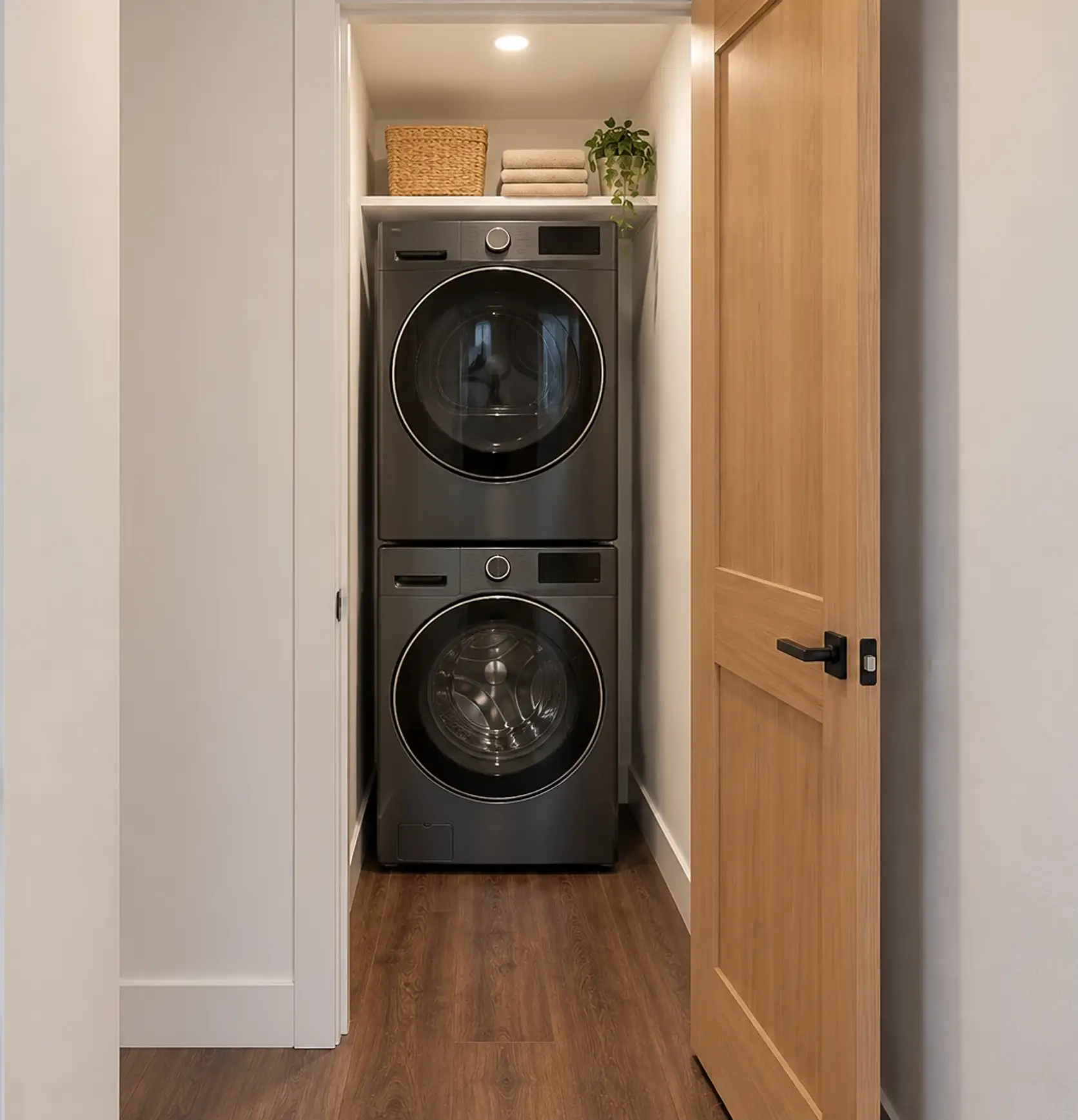 Rendering of the ADU 1200 laundry from the hall: a stacked graphite washer and dryer under a white shelf, through the open doorway, with the light-oak door standing open on the right.