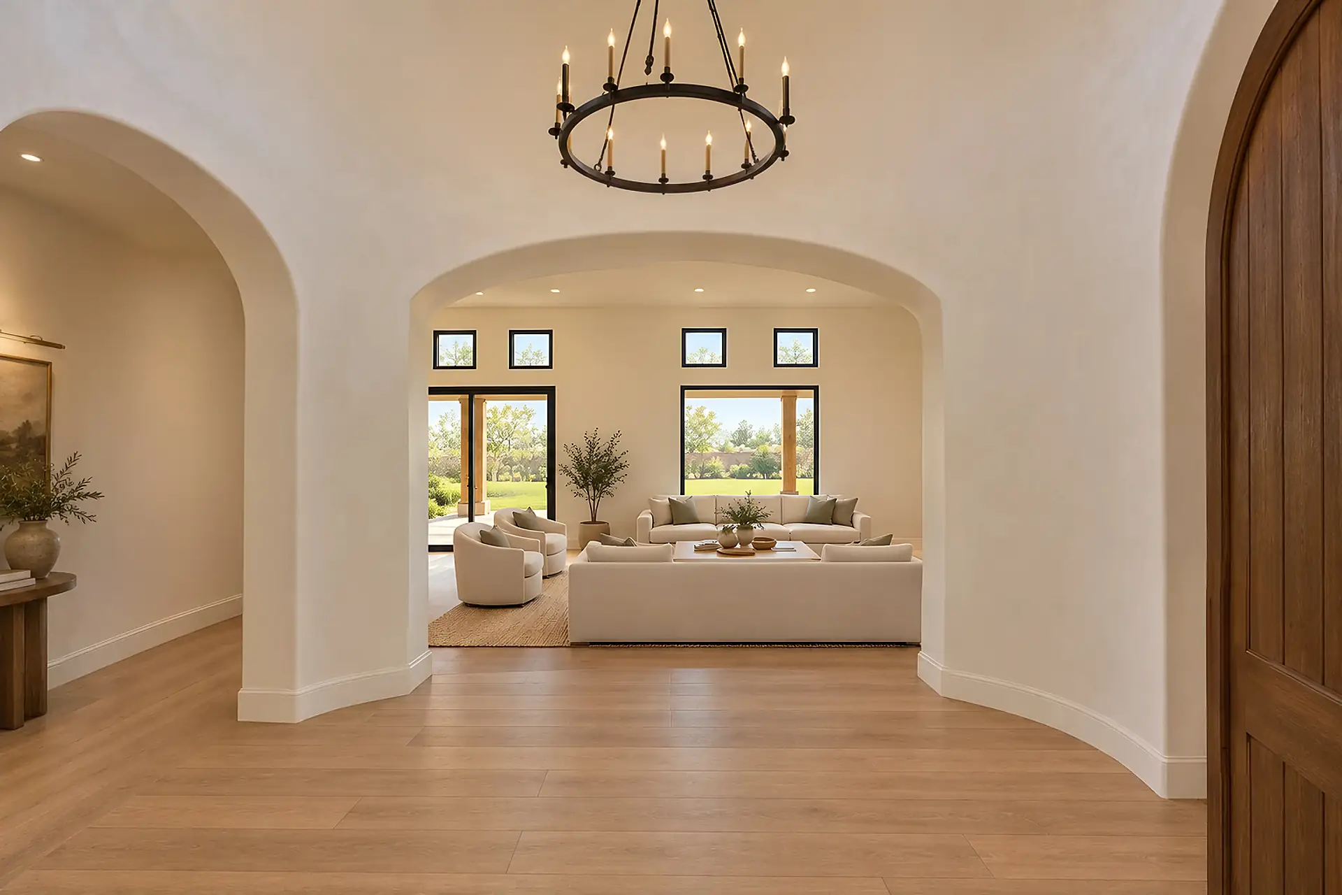 Rendering of the round entry rotunda from the front door: curved plaster walls under an iron chandelier, an oak floor, and wide arched openings into the great room ahead and the hall on the left, with the arched wood door to the garage at the right edge.